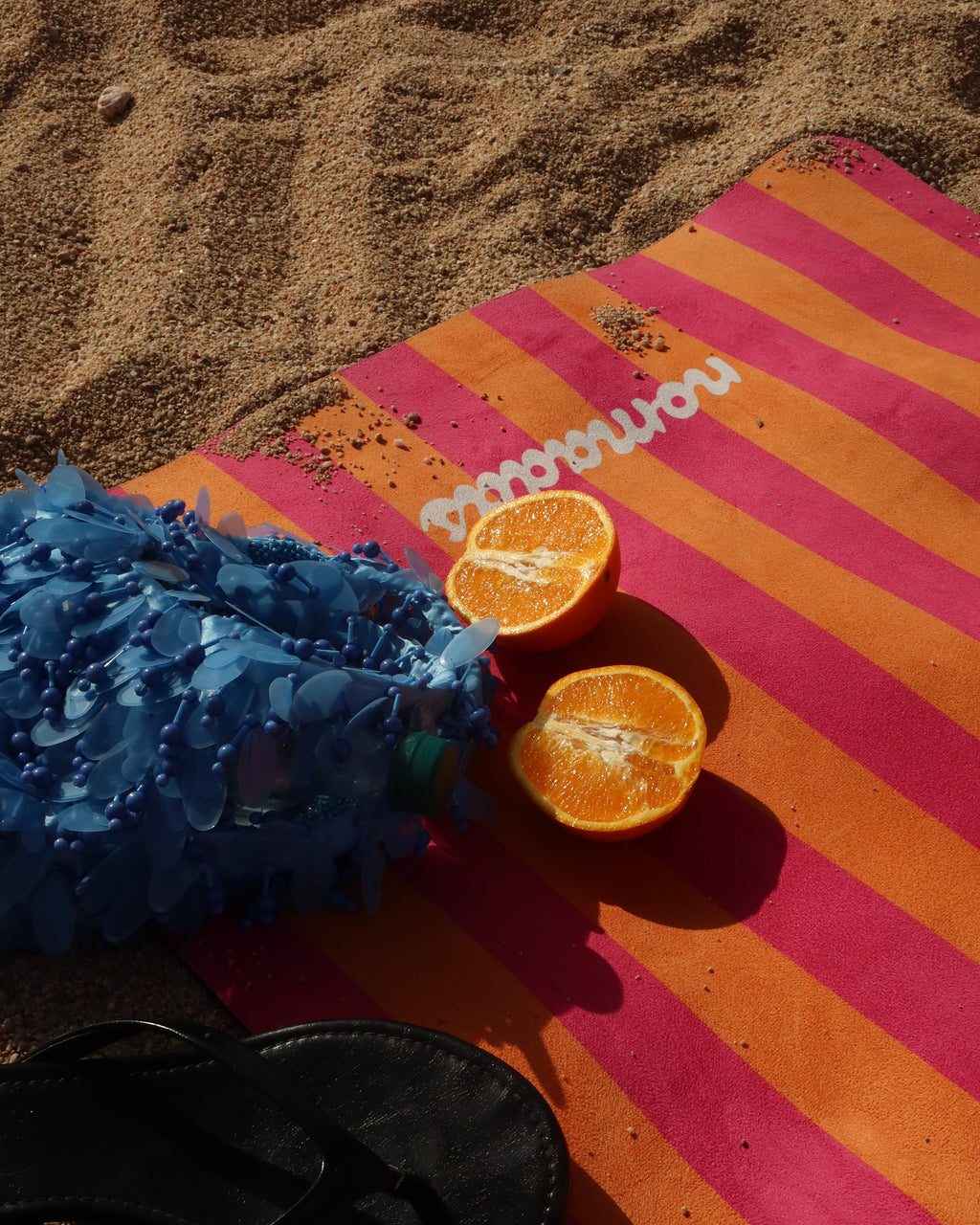 A top-down beach scene showing a pink and orange striped yoga mat on sand, alongside a blue ruffled beach umbrella, sliced oranges, a white straw, and a black flip-flop.